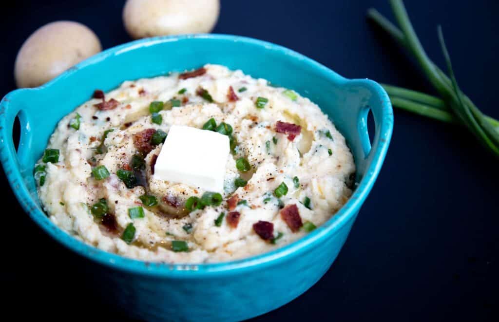 Cottage cheese mashed potatoes in a blue bowl topped with bacon, blue cheese, green onions, and butter. 