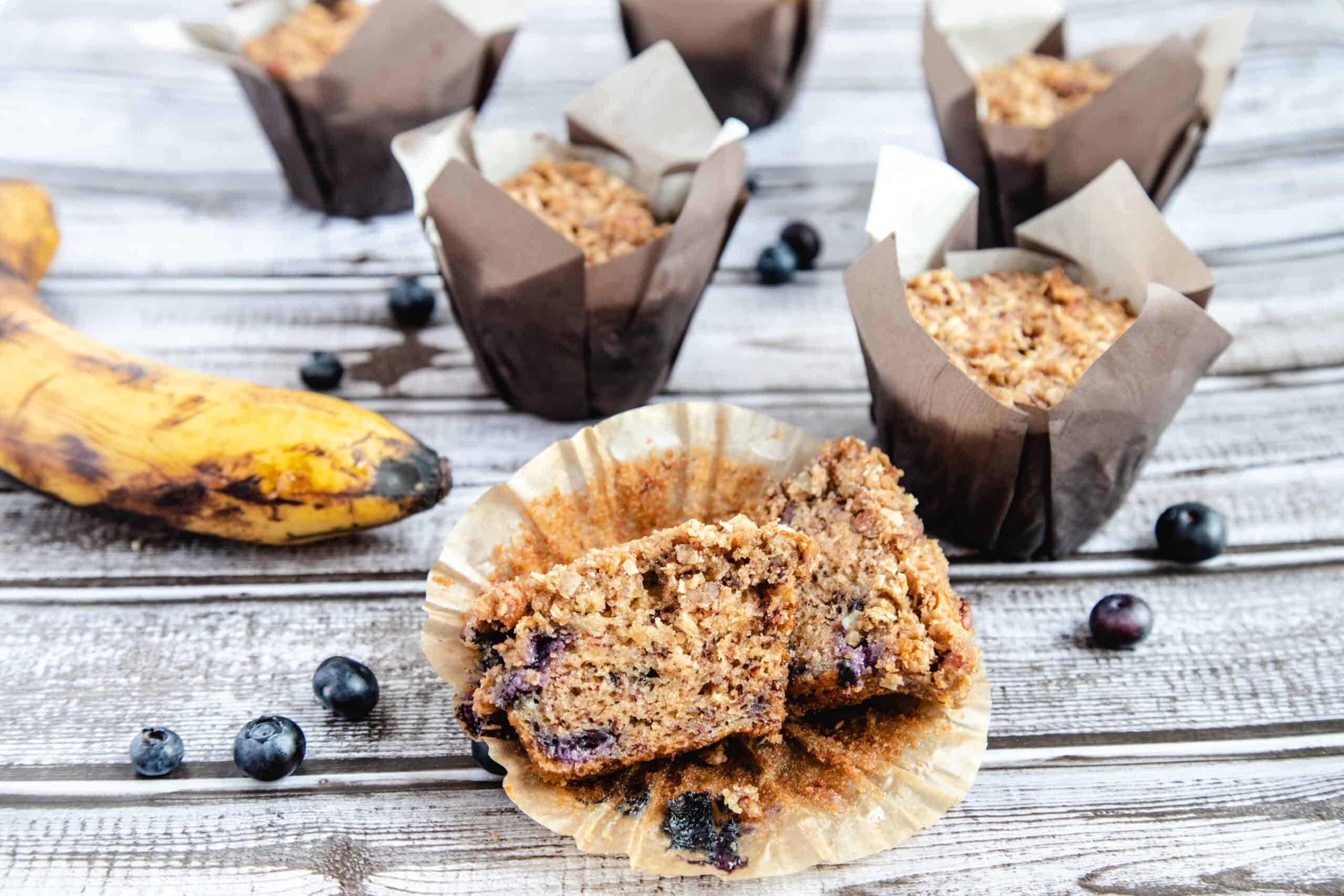 A blueberry, banana, and oatmeal muffin cut in half on a counter. There are full muffins in the background. 