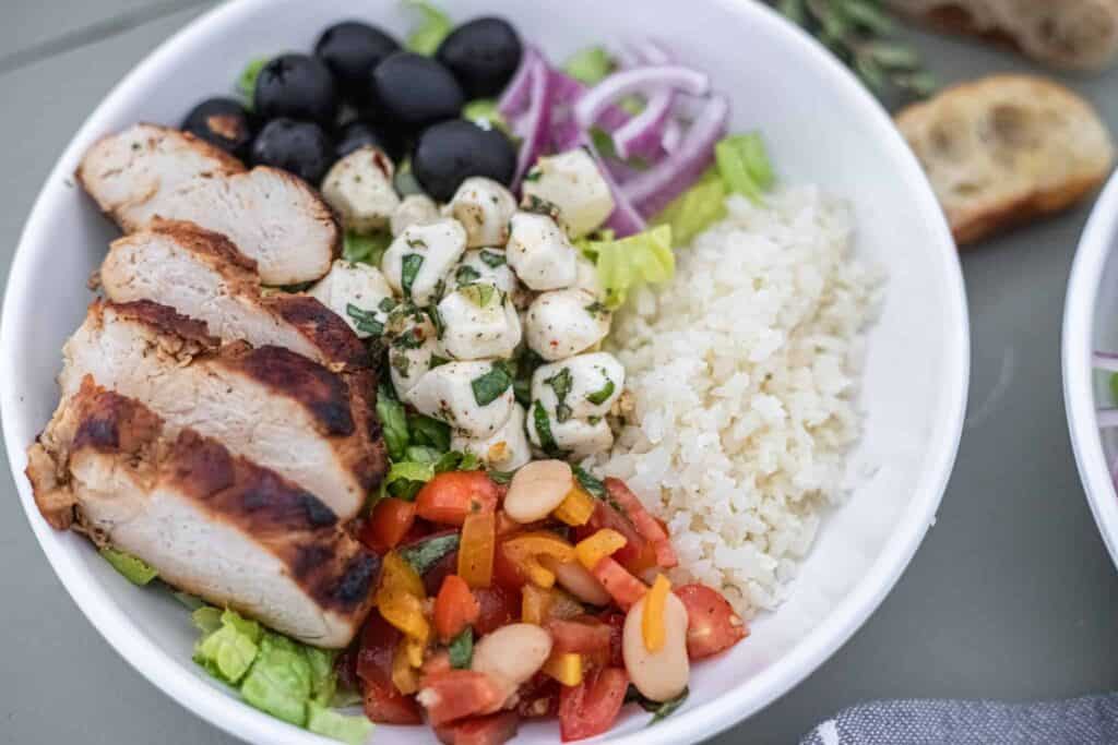 This is an up-close picture of a bowl with grilled chicken and bruschetta in a bowl with cauliflower rice, mozzarella pearls, black olives, romaine lettuce, and red onion. The Hangry Economist.