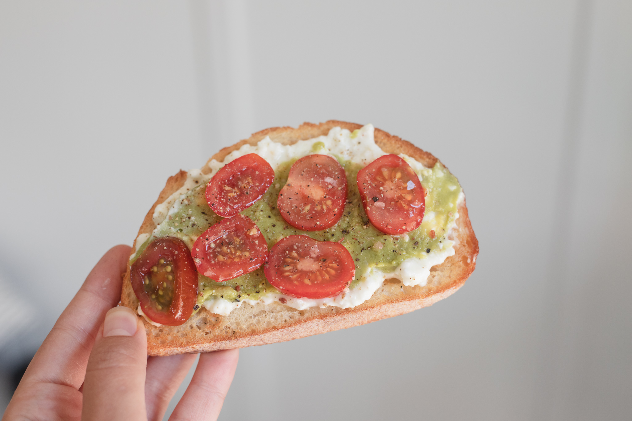 A hand holding up a piece of toast topped with cottage cheese, avocado, and tomatoes. 