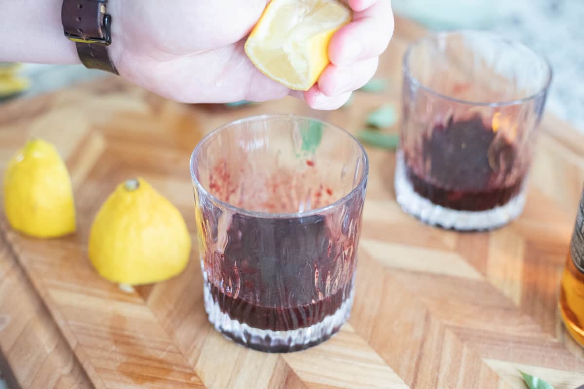 Lemon being squeezed into a cocktail glass filled with muddled blackberries and bourbon. 