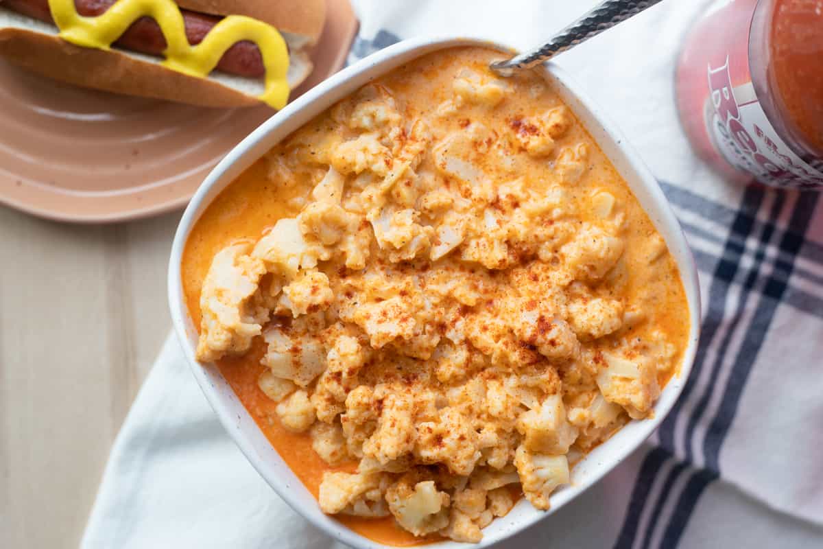 Buffalo cauliflower mac and cheese in a bowl next to a hot dog and a bottle of hot sauce.