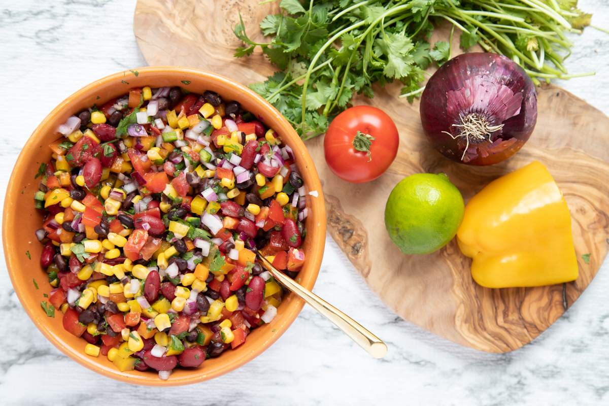 A pepper, red onion, tomato, lime, and cilantro on a cutting board next to a large bowl of bean and corn salsa. 