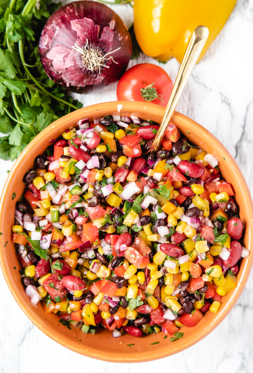 A large bowl of corn and bean salsa next to an onion, bell pepper, tomato, and bunch of cilantro. 