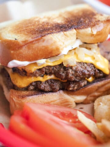 A grilled cheese burger in a basket with chips and tomato slices.
