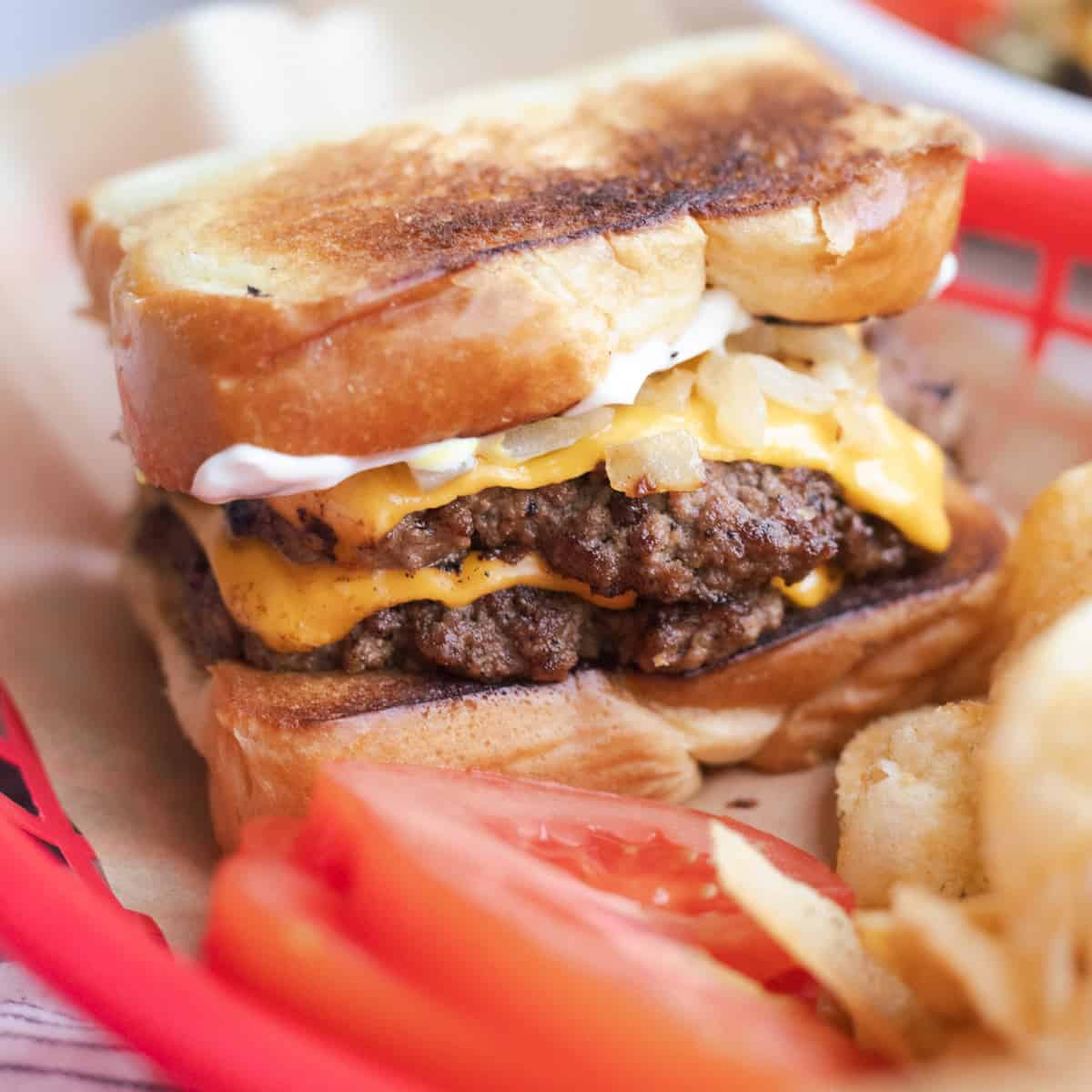 A grilled cheese burger in a basket with chips and tomato slices.