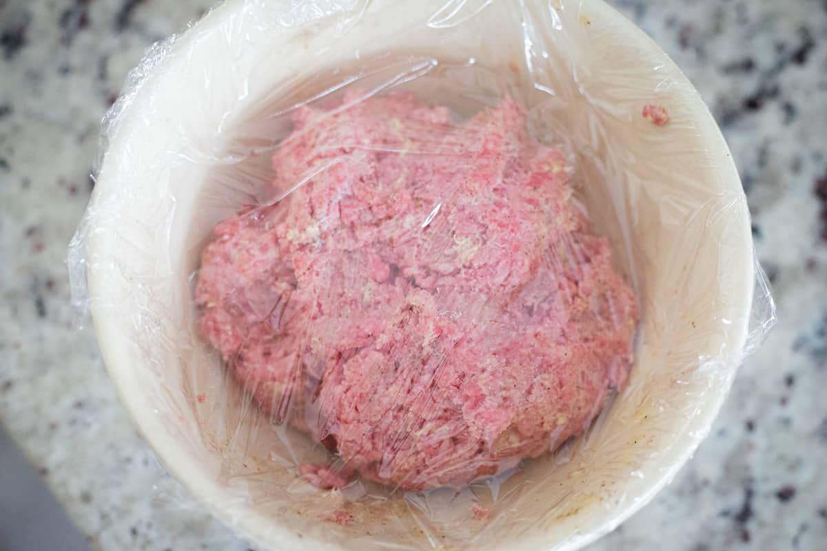Ground beef in a mixing bowl covered with plastic wrap. 