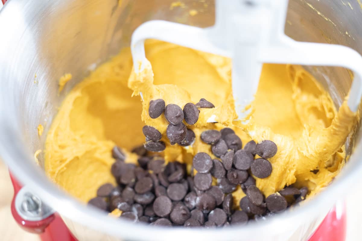 Chocolate chips being added to a pumpkin mixture in the bowl of a stand mixer. 