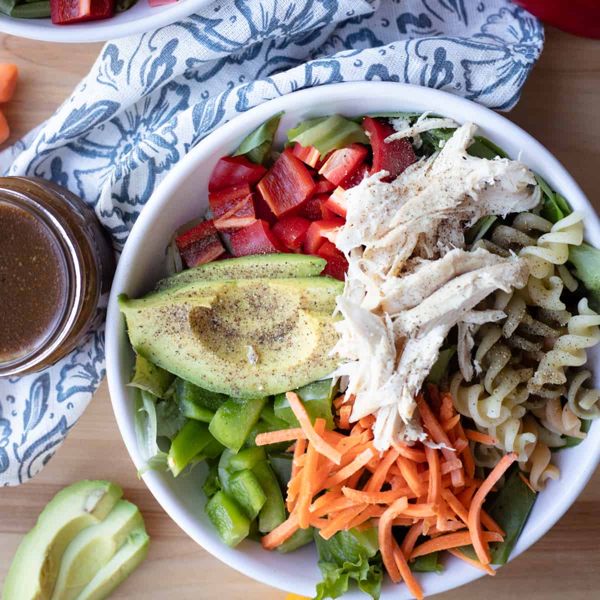 A bowl of rotisserie chicken garden salad with chicken, avocado, bell pepper, carrots, fusilli pasta, and salad dressing on the side.