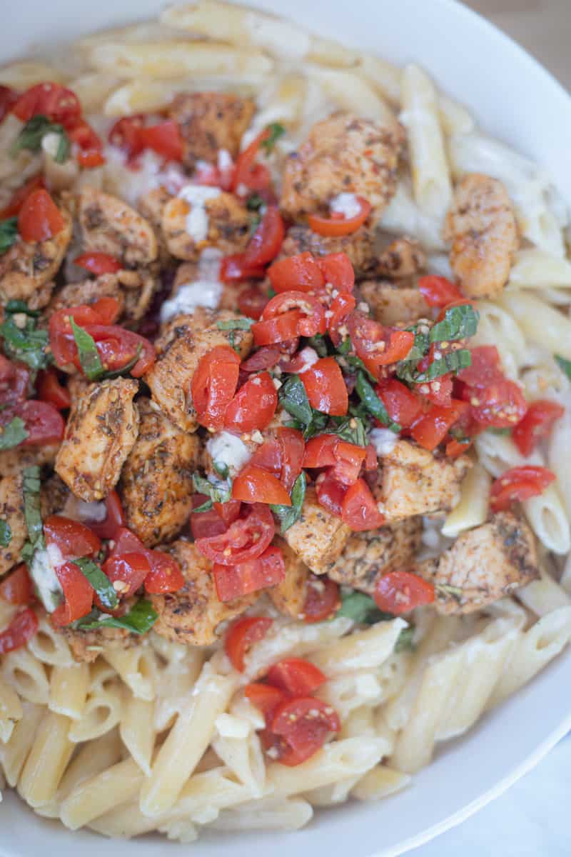 A serving bowl full of creamy three cheese penne topped with bruschetta tomatoes and chicken. 