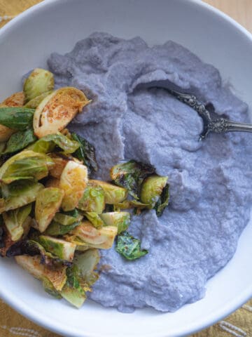 A bowl of purple cauliflower puree with a side of brussels sprouts.