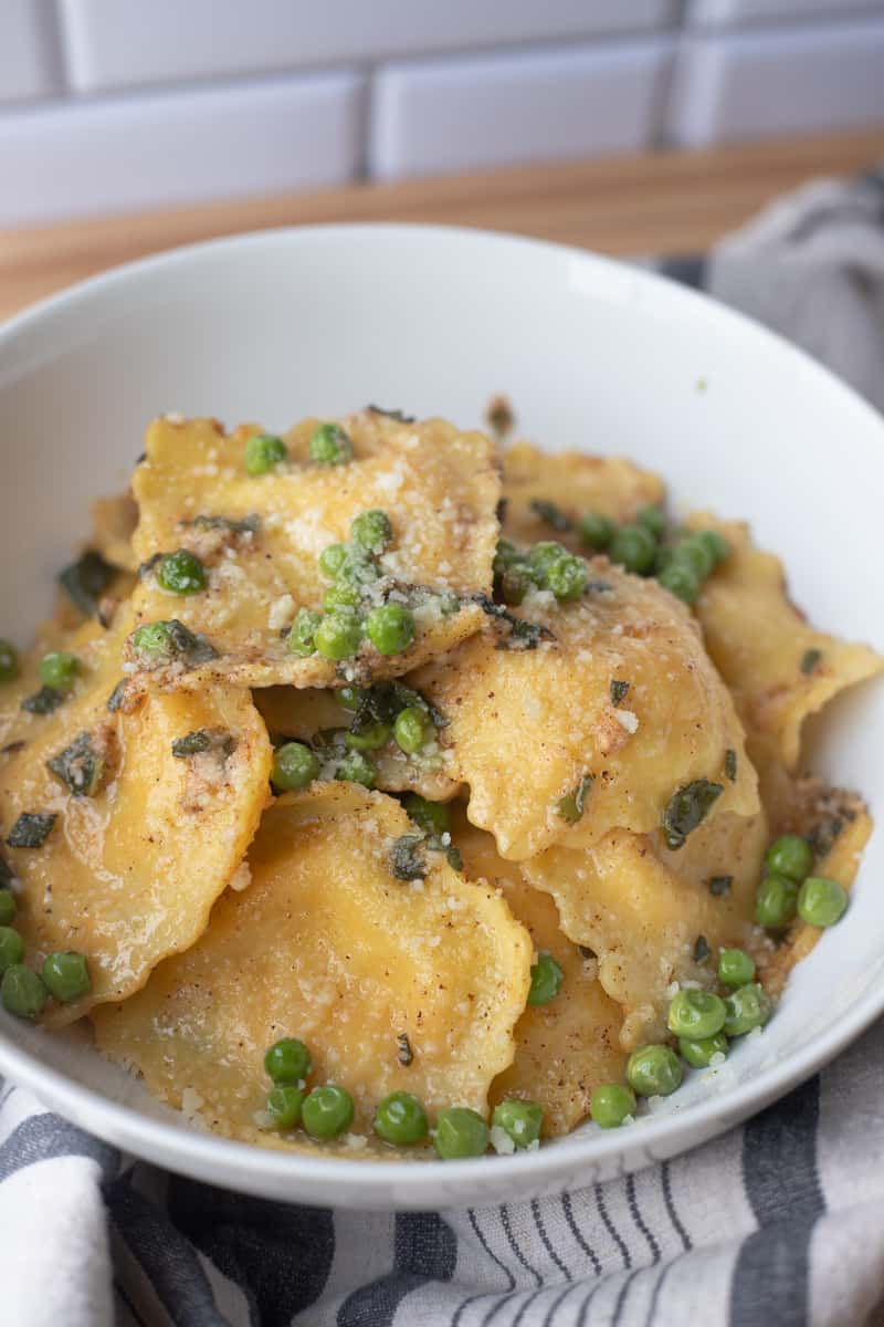 Brown butter sage ravioli in a bowl with peas and parmesan cheese. 