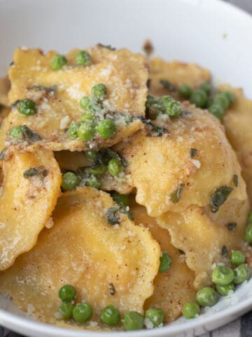 Brown butter sage ravioli in a bowl with peas and parmesan cheese.