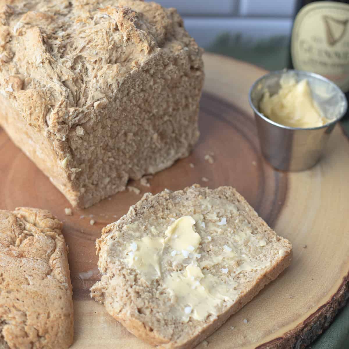 A slice of Guinness beer bread being slathered with butter with a loaf in the background. 