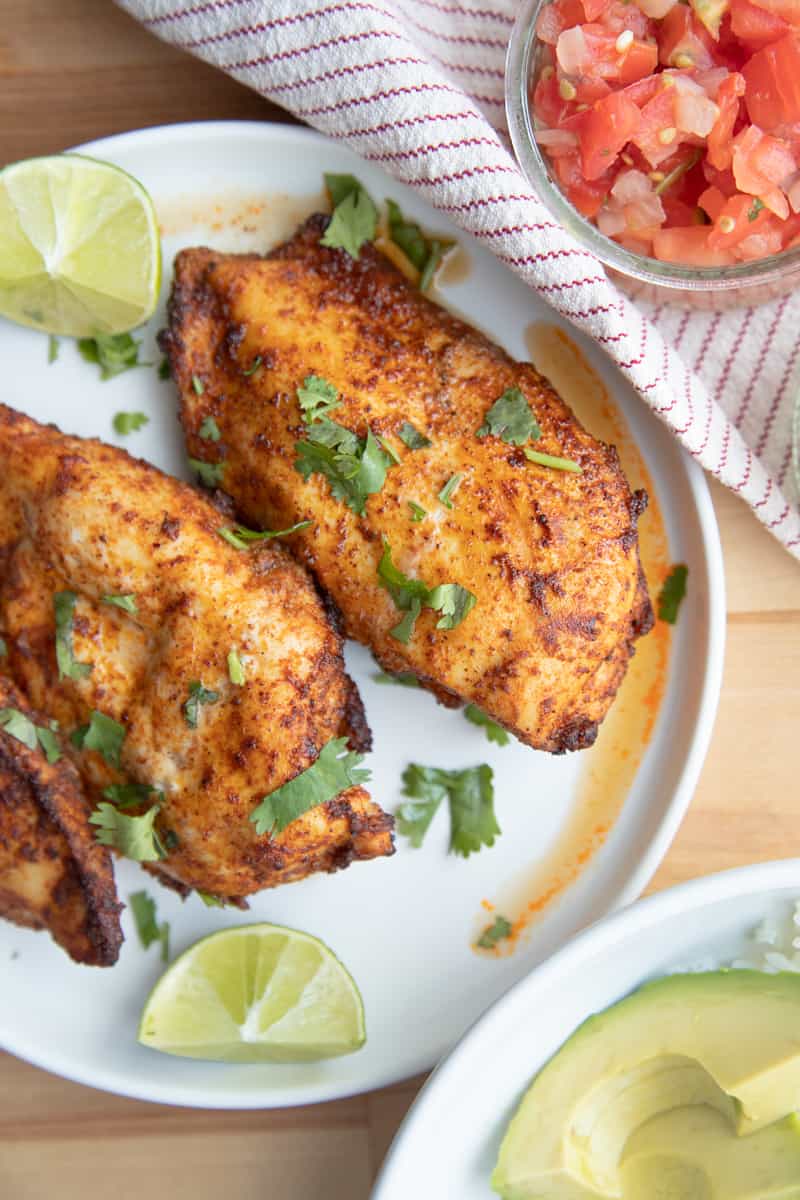 Two air fried blackened chicken breasts on a plate with limes and chopped cilantro. 