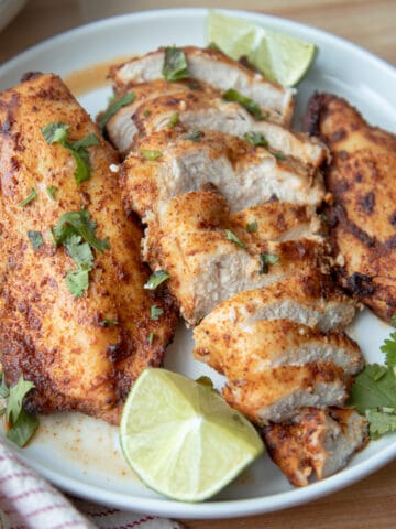 Three blackened chicken breasts on a plate garnished with limes and chopped cilantro. The middle chicken breast is sliced.