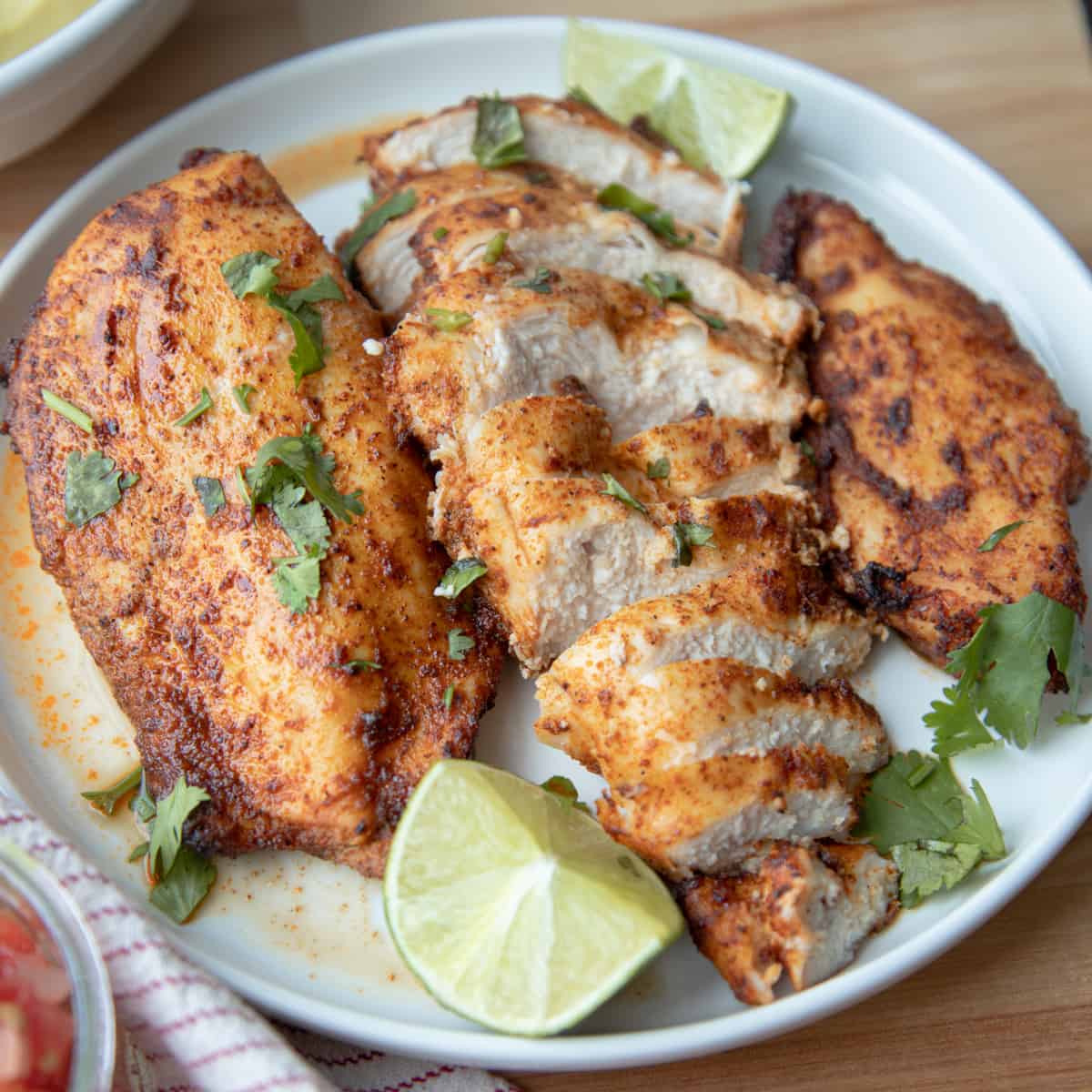 Three blackened chicken breasts on a plate garnished with limes and chopped cilantro. The middle chicken breast is sliced. 