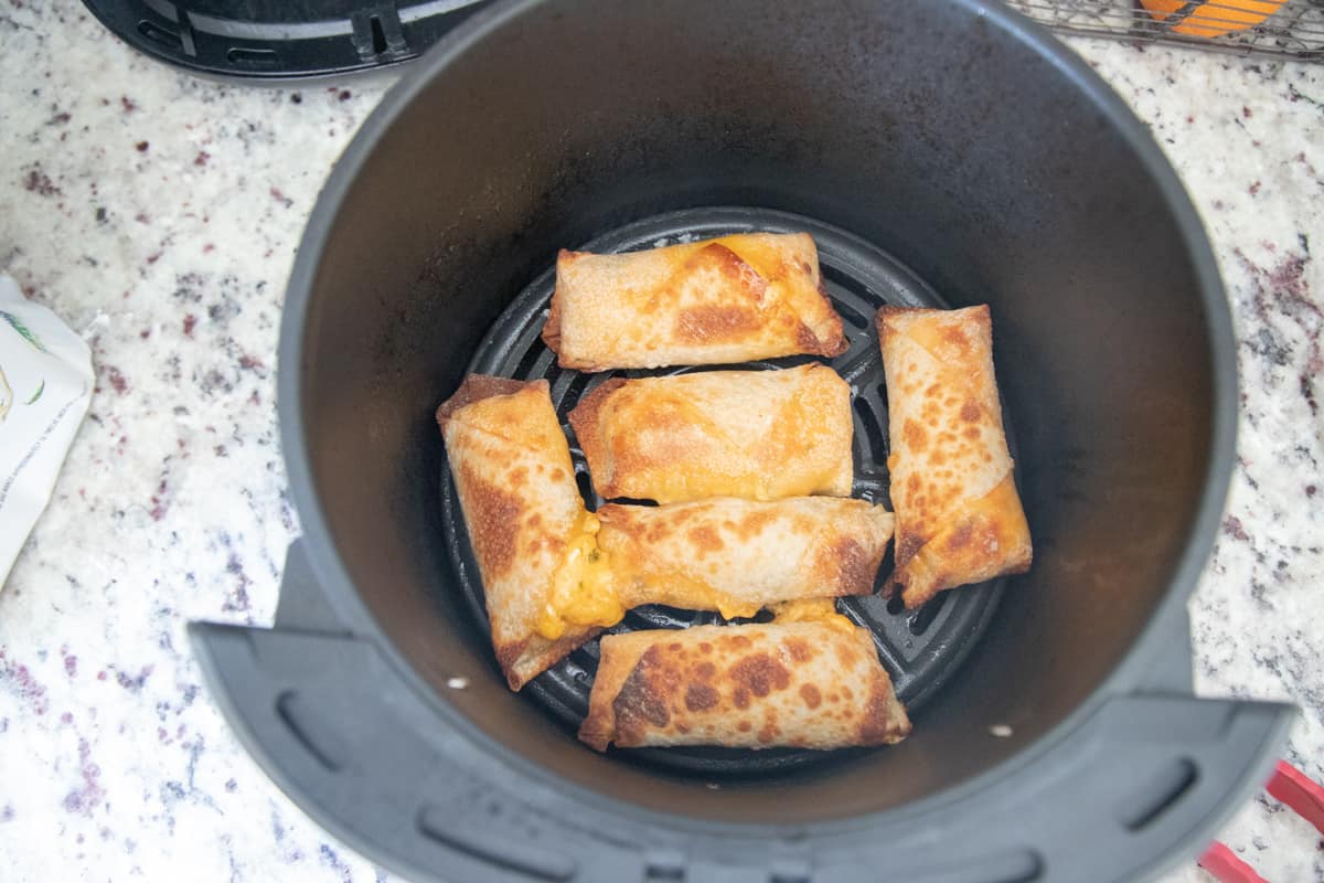 Six fully cooked buffalo chicken egg rolls in an air fryer basket. 