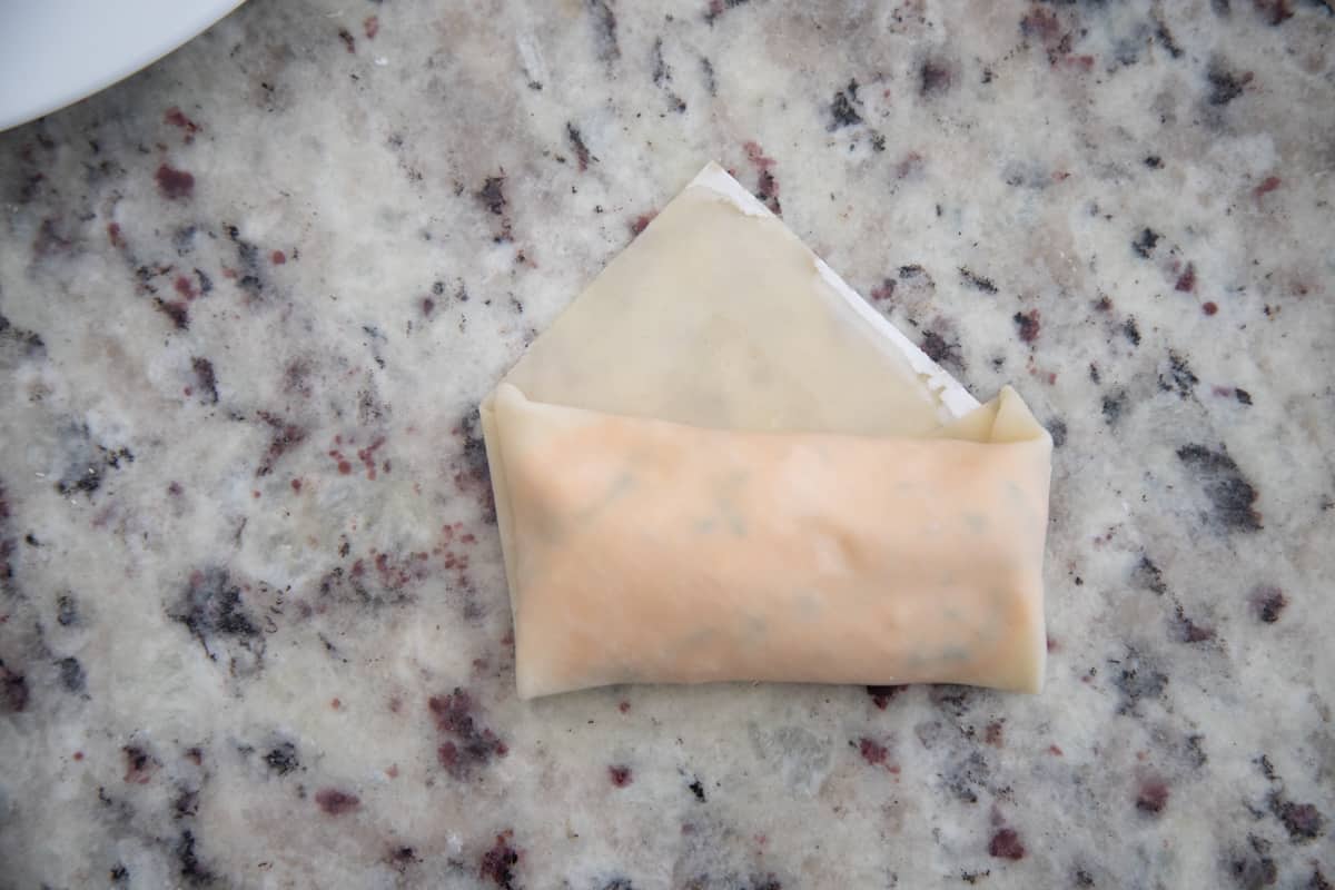 An egg roll wrapper with filling. The side corners and bottom corner are folded in, and the egg roll is mostly folded up, leaving a small triangle at the top. 