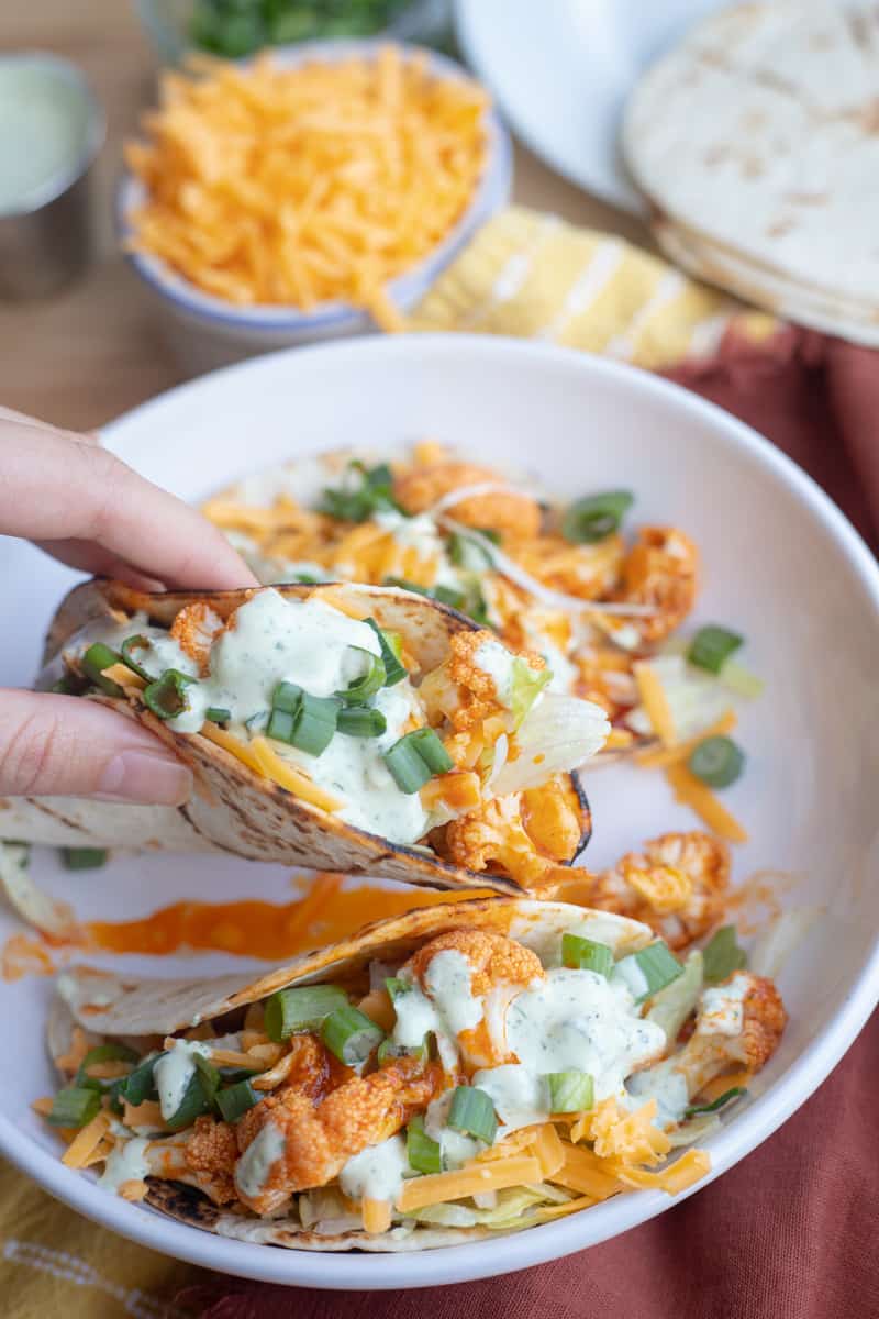 A hand holding a buffalo cauliflower taco topped with cilantro ranch, green onions, lettuce, and cheese. 