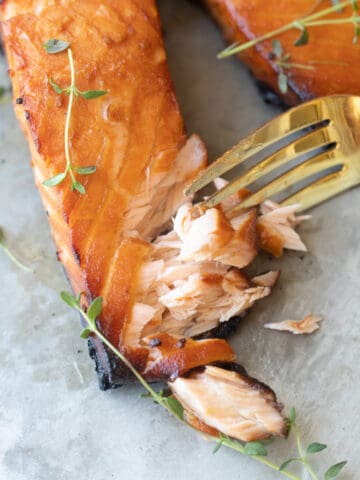 Two grilled salmon filets on a baking sheet topped with herbs. A fork is cutting into one of them.