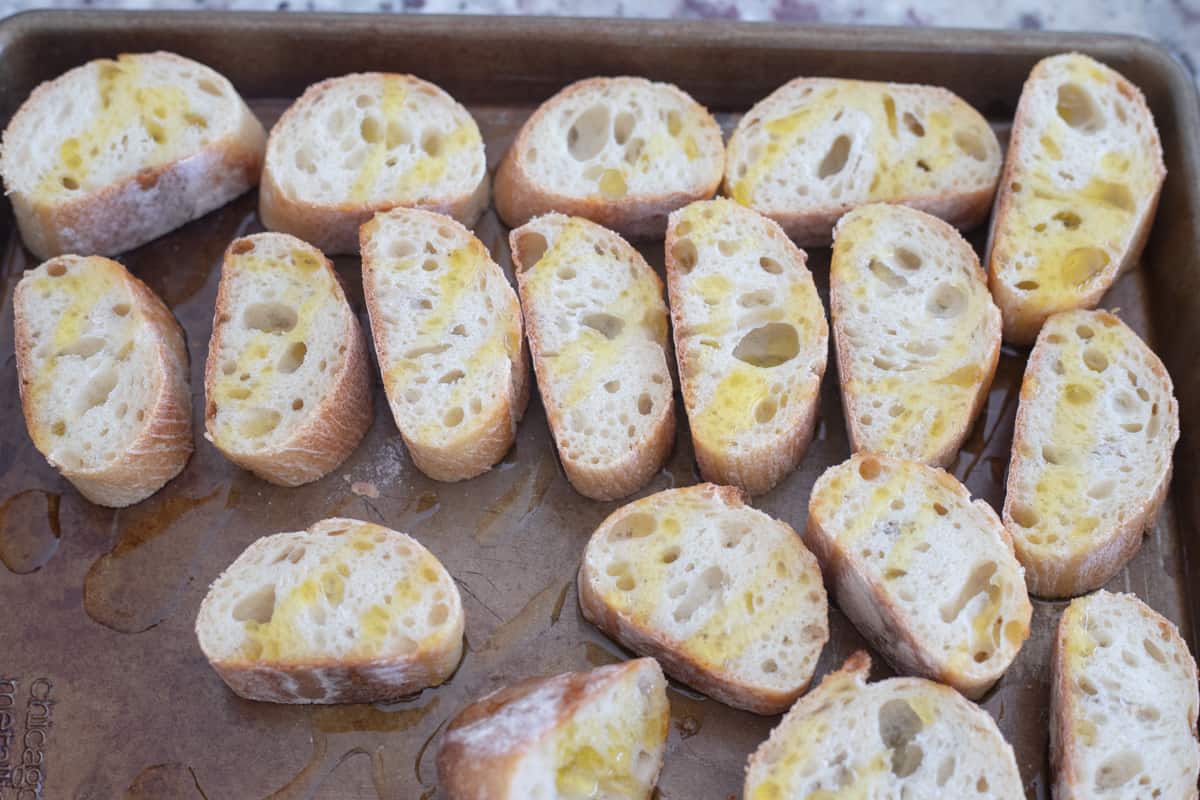 Slices of french bread on a sheet pan topped with olive oil. 