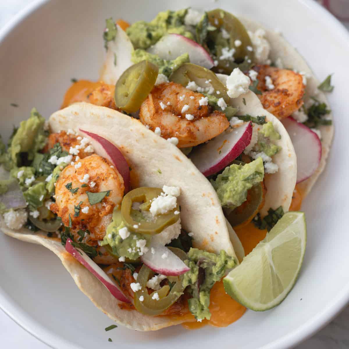 Three shrimp tacos on a serving plate topped with cheese, radishes, jalapeños, and cilantro.