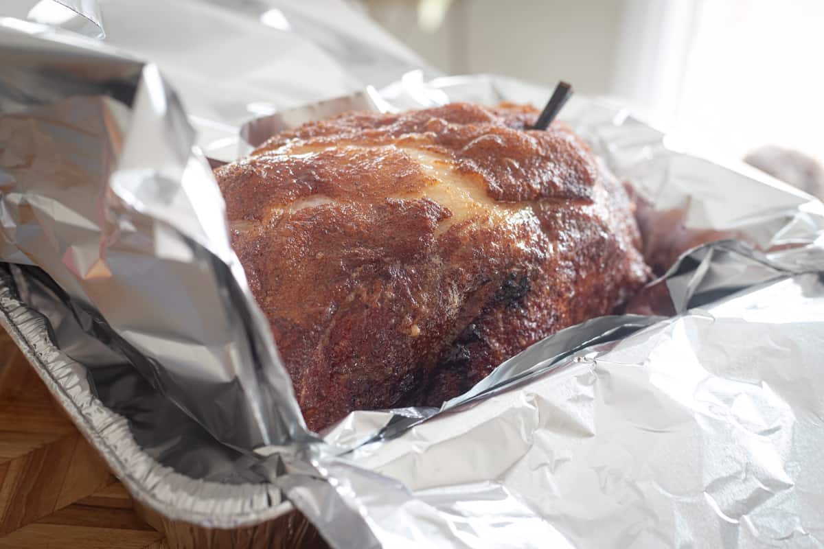 Pork butt wrapped in aluminum foil that's arranged cross-wise on top of an aluminum foil pan. 