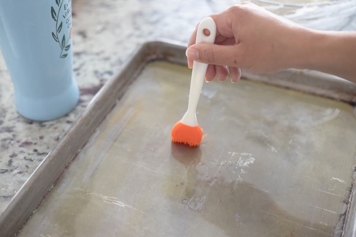 A hand oiling a sheet pan with olive oil and a silicone brush. 