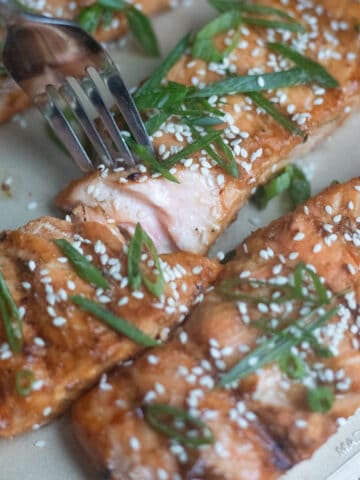 A grilled miso salmon fillet on a baking sheet with a fork breaking into it. It is garnished with green onions and sesame seeds.