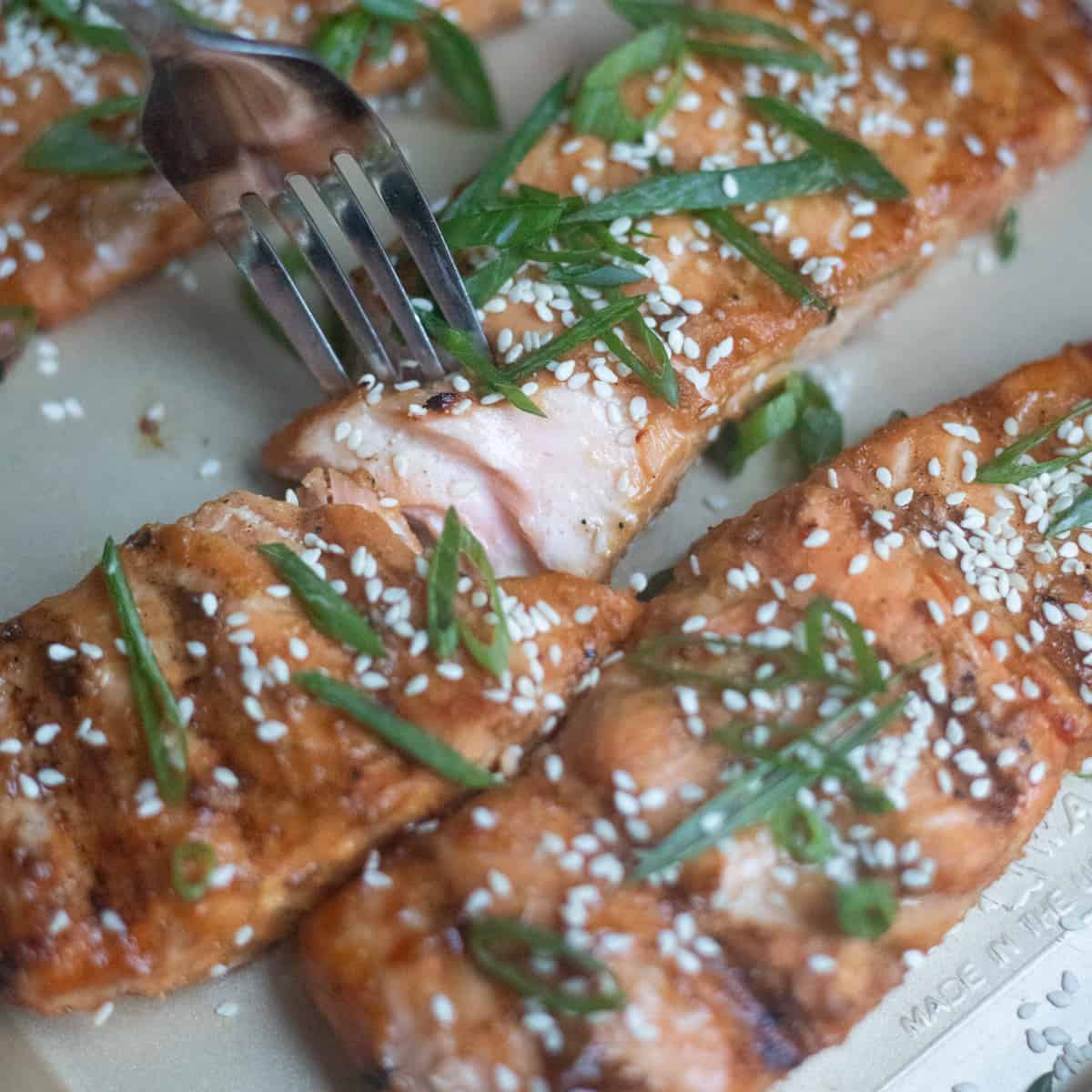 A salmon fillet on a baking sheet with a fork breaking into it. It is garnished with green onions and sesame seeds.