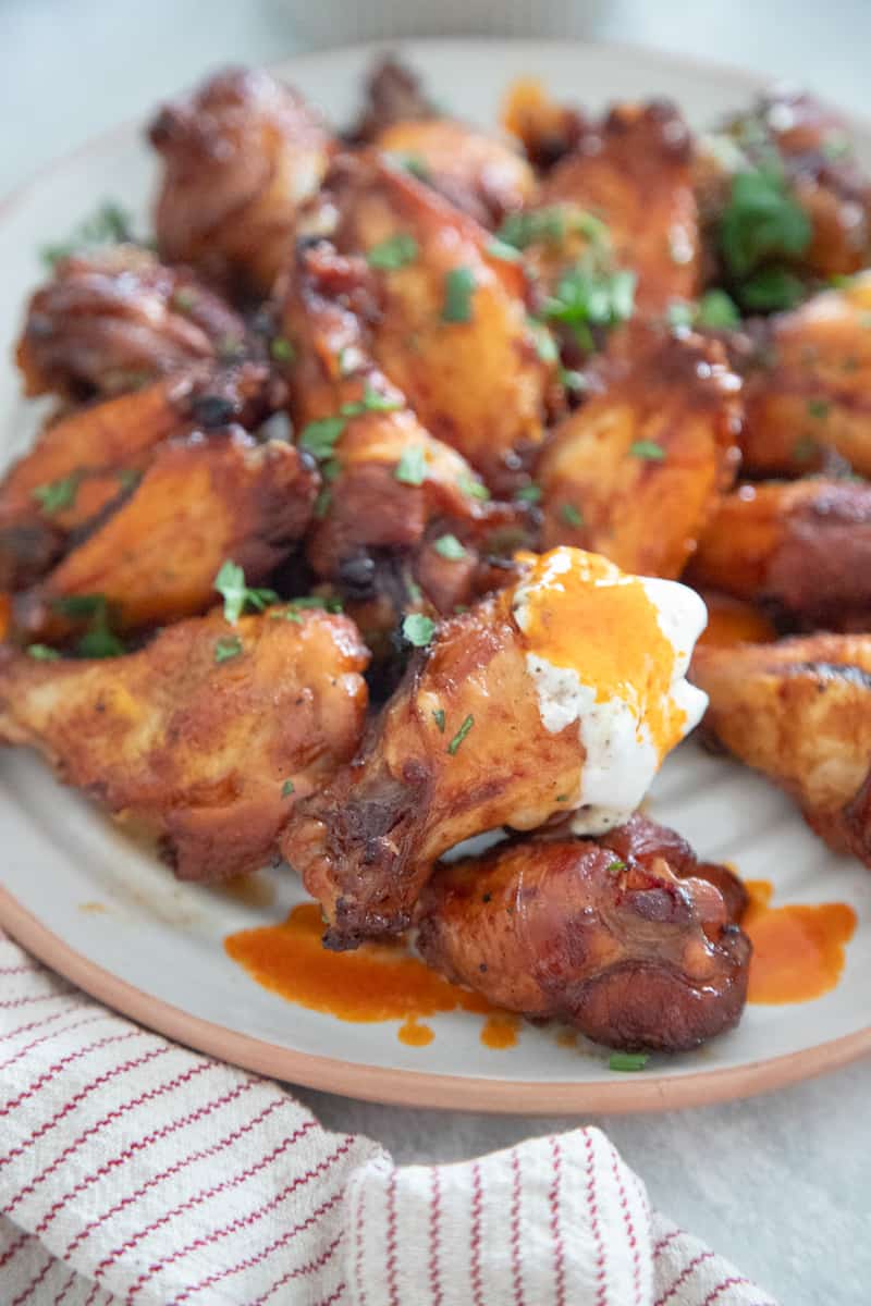 Several chicken wings on a plate. They're coated in hot sauce. One wing on the edge of the plate is dipped in ranch dressing. 