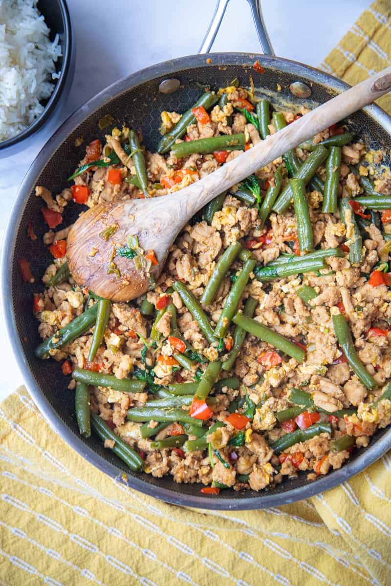 A pan of ground chicken stir fry with a wooden spoon resting on top. Green beans, red peppers, and ground chicken are visible. 