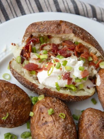 A plate full of baked potatoes. One is topped with sour cream, bacon, cheese, and green onions.