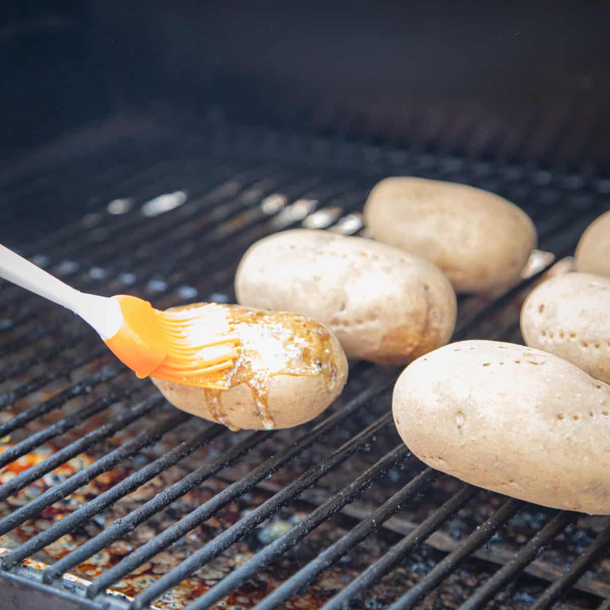 Butter being spread on a baked potato with a basting brush on a Traeger grill. 