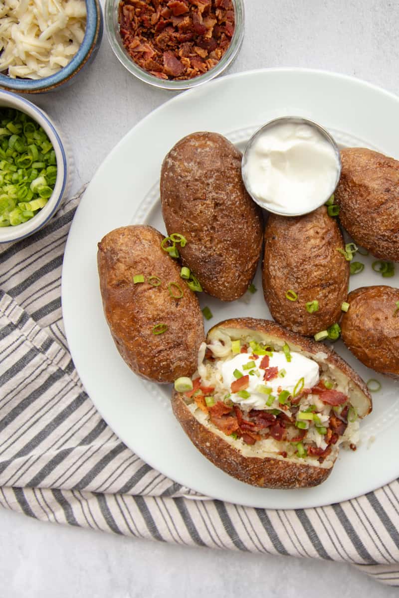 A plate full of baked potatoes. One is topped with sour cream, bacon, cheese, and green onions. There is a dish of sour cream on the plate and bowls of toppings on the side. 