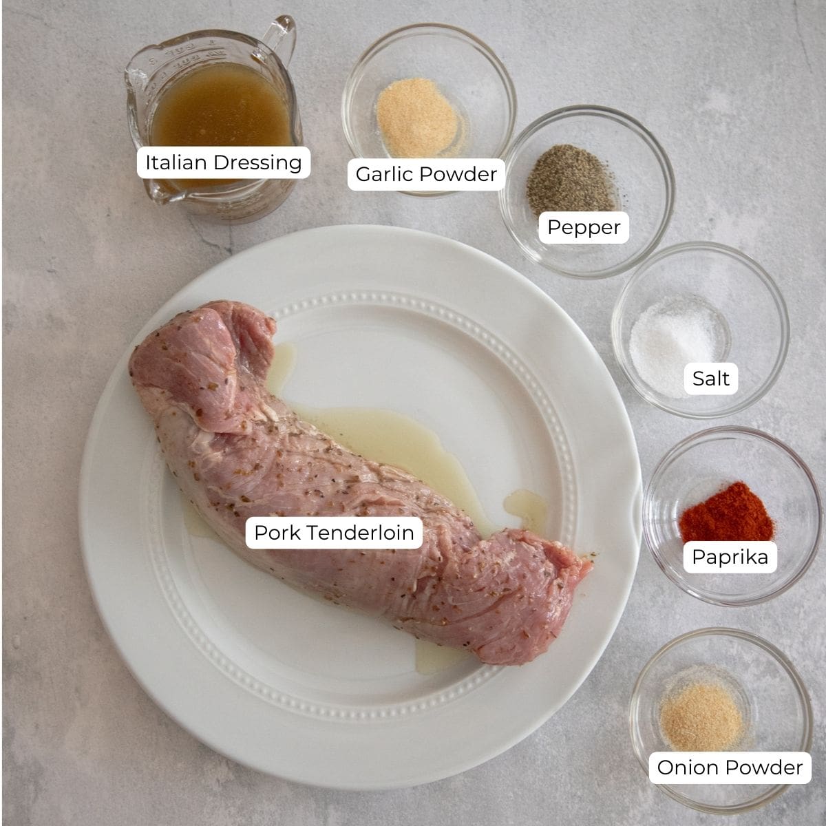 Ingredients for Traeger pork tenderloin on a countertop. Pictured is spices, Italian salad dressing, and a raw pork tenderloin. 