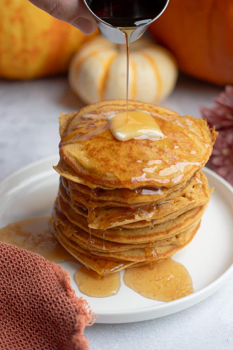 A stack of gluten free pumpkin pancakes topped with butter. Syrup is being poured on top and there are pumpkins in the background. 