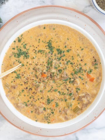 A bowl of hamburger rice soup topped with a green garnish. There is a spoon in the bowl and the soup is dark yellow in color.
