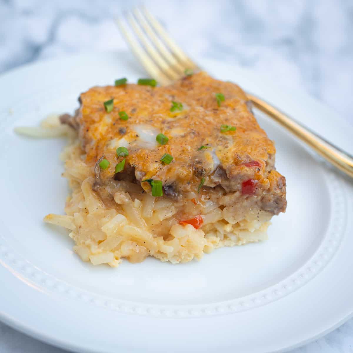 A square of egg and sausage gravy casserole on a plate. A fork is resting on the side. The casserole is orange in color and topped with chopped chives.