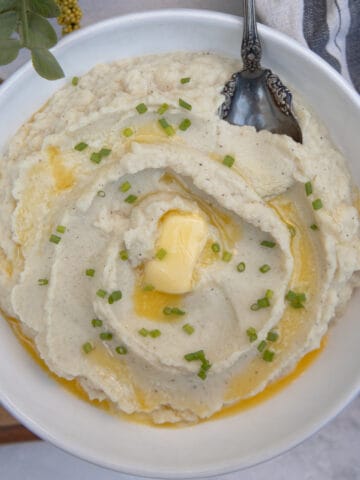 A white bowl of mashed cauliflower topped with melted butter and chopped chives.