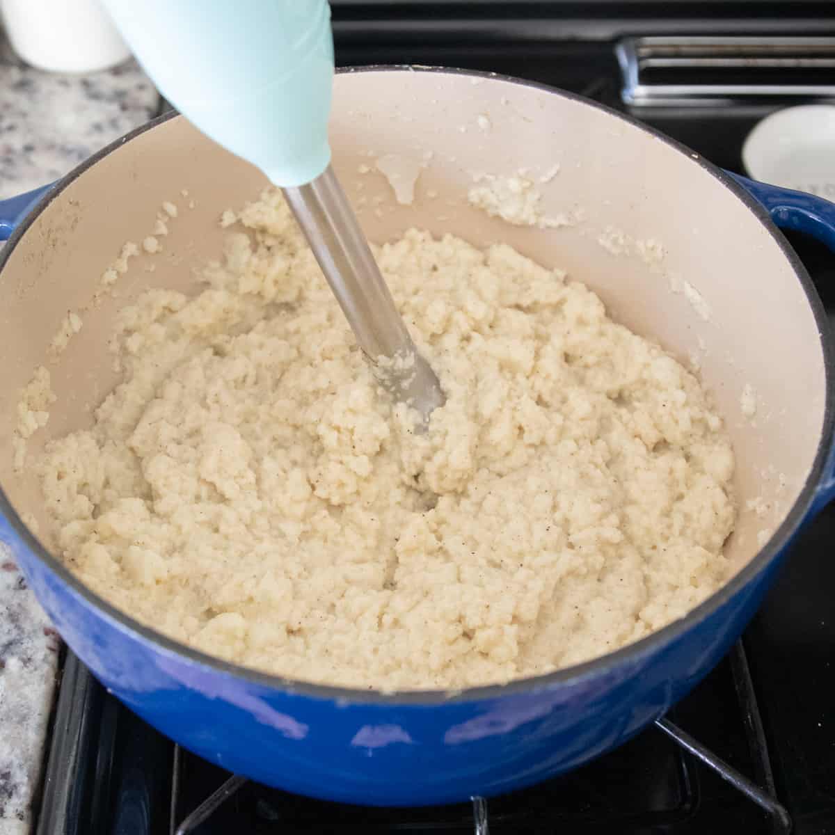 Cauliflower being blended  with an immersion blender in a blue dutch oven. 