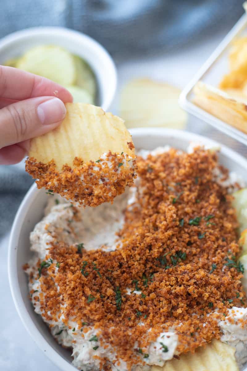A chip being dipped into fried pickle dip that's garnished with toasted panko breadcrumbs and fresh dill. 