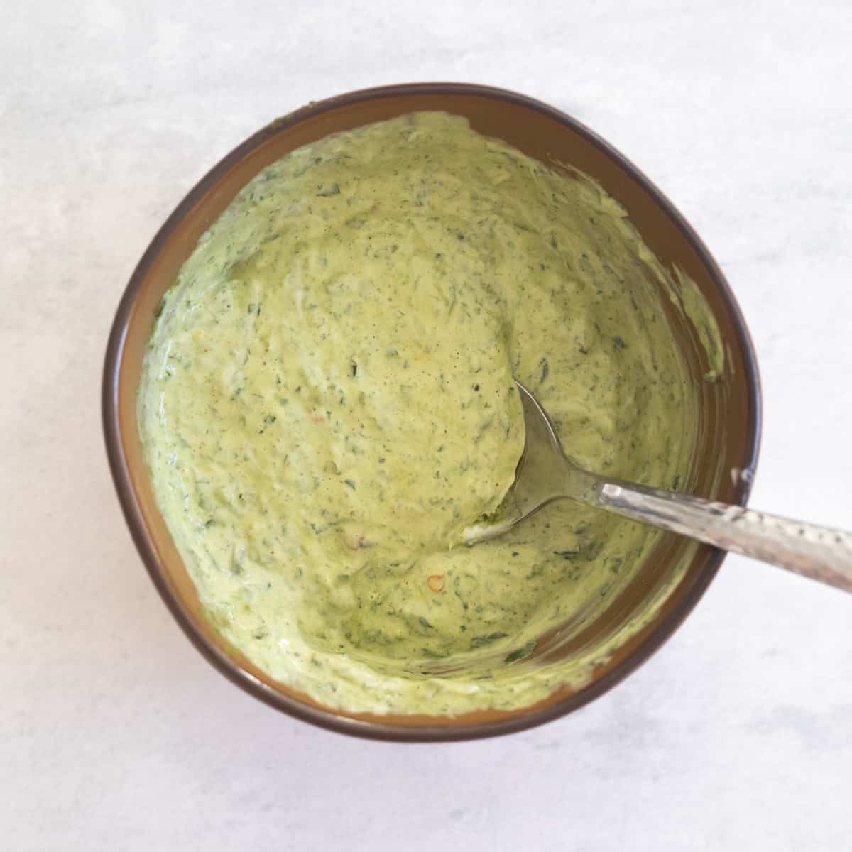 Pesto mixed with mayonnaise in a brown bowl. A spoon is resting in the mixture. It's light green in color with dark green specs. 