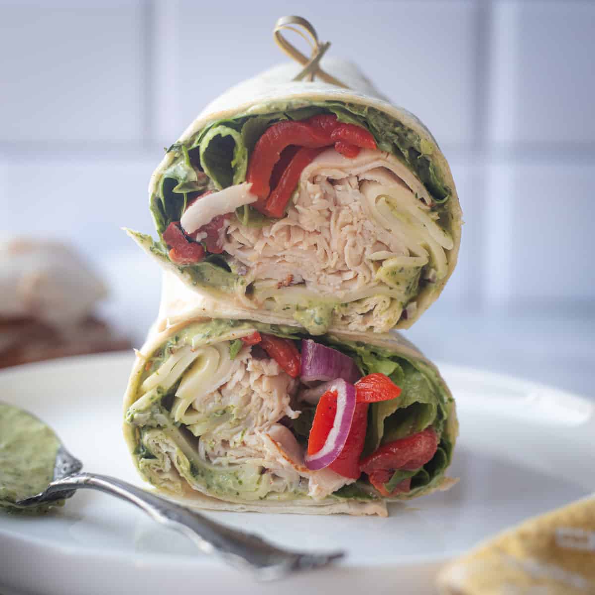 Two halves of a pesto turkey wrap on a plate. They're stacked on top of each other and each one is stuffed with turkey, roasted red peppers, lettuce, onions, and pesto. 