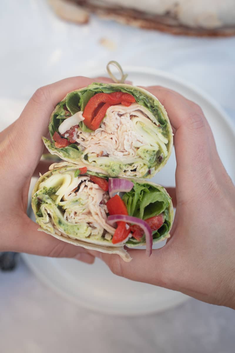 Hands holding two halves of a pesto turkey wrap stuffed with turkey, lettuce, roasted red peppers, onions, and cheese. 