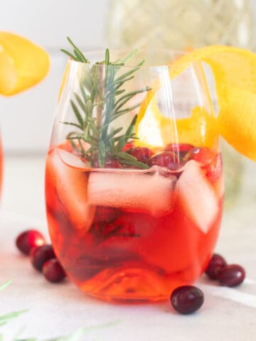 A stemless wine glass filled with a winter aperol spritz. It's an orange-red drink, garnished with an orange twist, rosemary, and fresh cranberries.