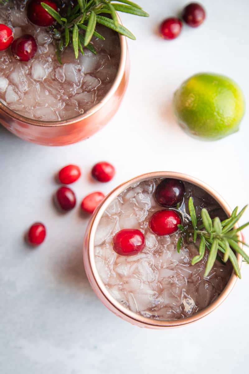 Two Christmas Moscow mules filed with ice on a countertop. They are garnished with fresh cranberries and rosemary. There is a lime resting to the side and several fresh cranberries. 