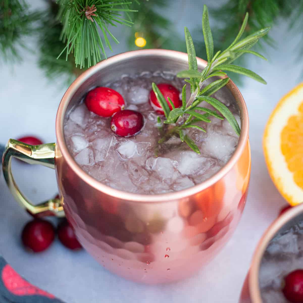 A Christmas Moscow mule filed with ice on a countertop. It is garnished with fresh cranberries and rosemary. There is an orange resting to the side and several fresh cranberries.