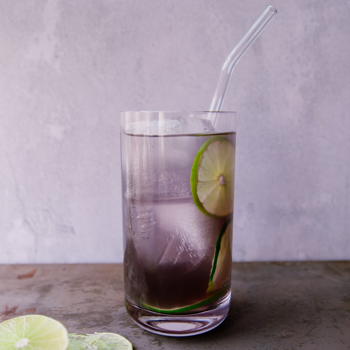 A purple rain cocktail in a tall cocktail glass. It's light purple in color and garnished with slices of lime. There is a clear straw in the glass.